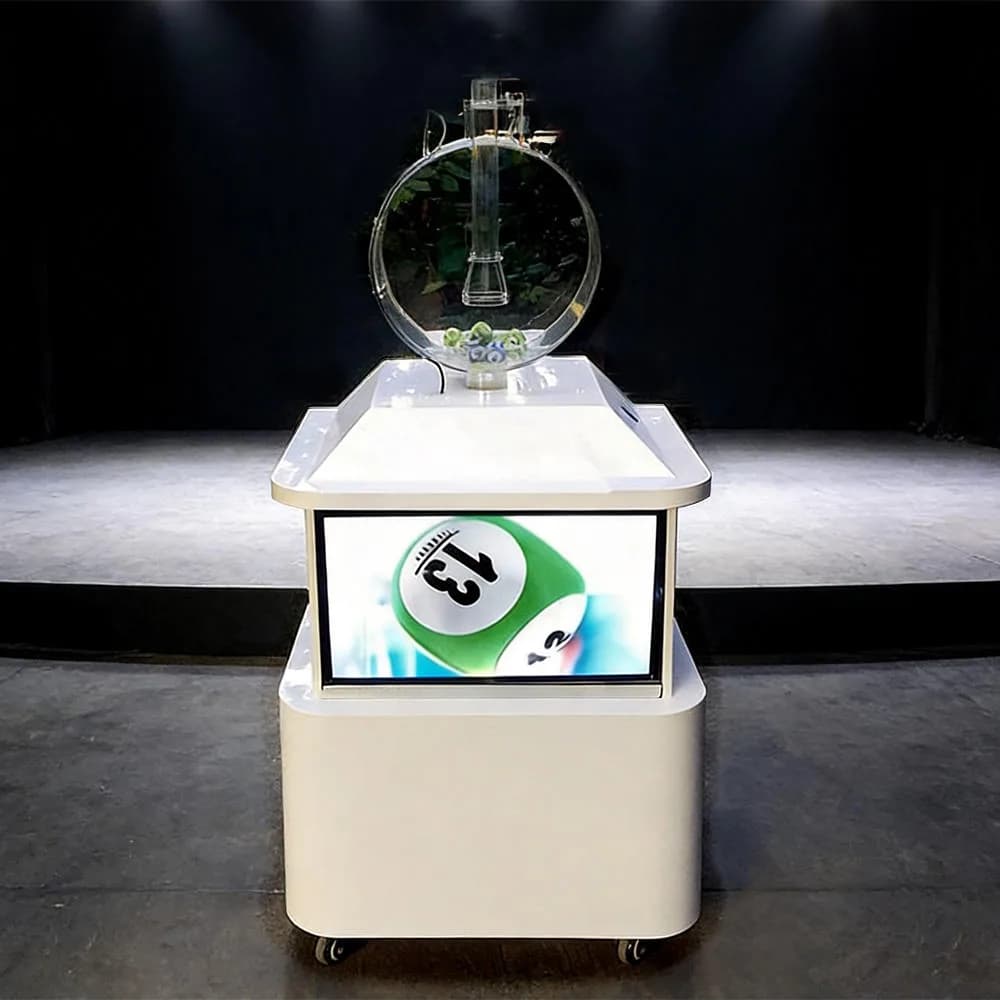Front view of a white single-cylinder pick 1 lottery machine with a TV screen showing a green number 31 ball, standing on a stage